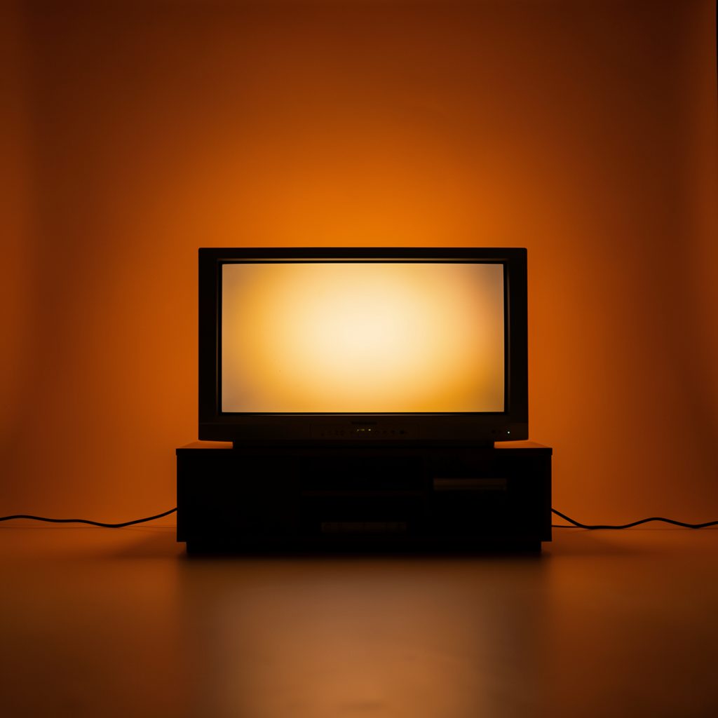 A dark cinematic wide shot of a glowing television screen in a dim room with warm amber light spilling across the floor, studio photograph on a seamless backdrop
